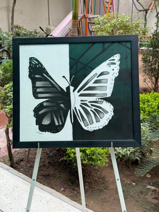 Black and white butterfly painting on canvas, divided into two contrasting halves – one side dark with a white butterfly pattern and the other side light with a black butterfly pattern, displayed outdoors on an easel.