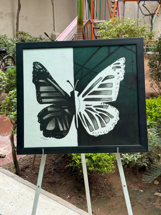Black and white butterfly painting on canvas, divided into two contrasting halves – one side dark with a white butterfly pattern and the other side light with a black butterfly pattern, displayed outdoors on an easel.